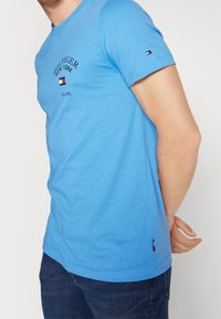 Muž v světle modrém tričku Tommy Hilfiger a tmavě modrých džínách, se sepnutýma rukama za zády.