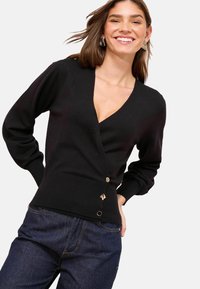 Femeie zâmbitoare cu păr brunet până la umeri, îmbrăcată cu un cardigan negru cu decolteu în V și trei butoane decorative, purtând blugi de culoare albastru închis.