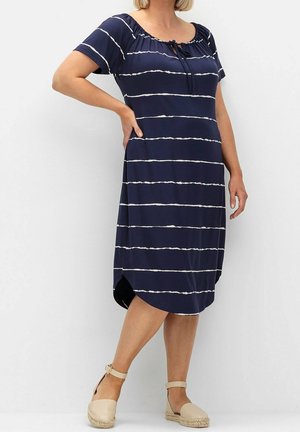 Frau in marineblauem Kurzarmkleid mit weißen horizontalen Streifen und beigen Espadrille-Pumps mit Knöchelriemen, steht mit einer Hand in der Hüfte.