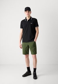Polo noir avec des bordures blanches, shorts verts, baskets noires et casquette noire. Le mannequin se tient avec une main dans la poche du short.