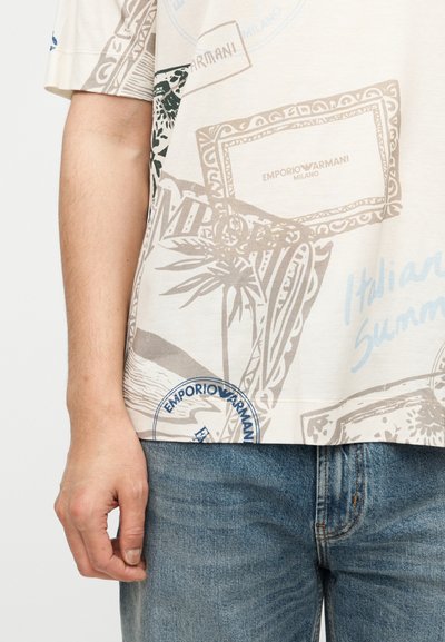 Κοντινό πλάνο στον κορμό ενός ατόμου που φοράει ένα μπεζ μπλουζάκι με τύπωμα «Emporio Armani» και μπλε τζιν, με το αριστερό χέρι χαλαρό στο πλάι.