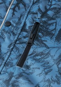 Niebieska tkanina z czarnym abstrakcyjnym wzorem, z zamykaną kieszenią na zamek i detalem w postaci guzika. Gładka tekstura z kontrastowym przeszyciem.