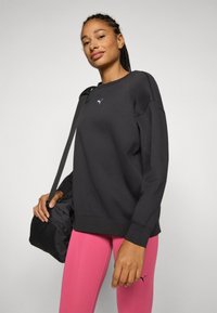 Sudadera negra con cuello redondo y corte holgado, combinada con leggings rosa. Presenta un pequeño logo blanco en el pecho.
