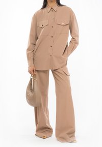 Femme portant une chemise beige à boutons et un pantalon large assorti, tenant un sac à main beige avec une bandoulière en chaîne dorée.