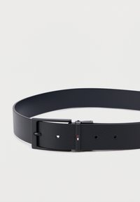 Černý kožený pásek s texturovaným povrchem, obdélníkovou kovovou sponou a pěti otvory pro nastavení velikosti. Jemné červené a bílé akcenty.