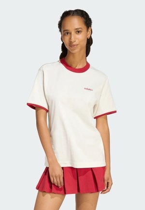 Femme aux cheveux tressés portant un t-shirt blanc avec bordure rouge et une jupe plissée rouge assortie sur un fond clair uni.
