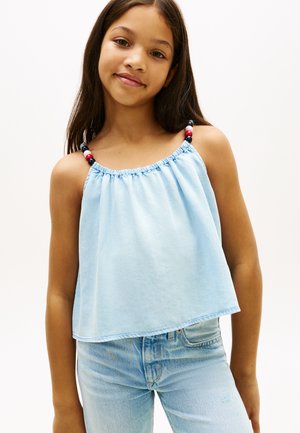 Ung pige iført en lyseblå ærmeløs top med perlebesatte stropper og lyseblå denimjeans, stående foran en ensfarvet baggrund.