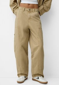 Pantalones cargo de pernera ancha en caqui con grandes bolsillos laterales, una tela texturizada y cierre de botón. Combinados con zapatillas blancas que tienen acentos negros.
