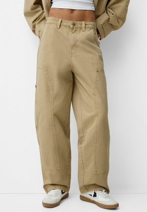 Pantalons cargo larges en kaki avec de grandes poches latérales, un tissu texturé et une fermeture à bouton. Associés à des baskets blanches avec des accents noirs.