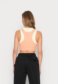 Sportowy biustonosz w jasnym brzoskwiniowym kolorze z kremowymi akcentami, z plecami w stylu racerback, teksturowanym materiałem i dopasowanym krojem. Noszony z czarnymi spodniami.