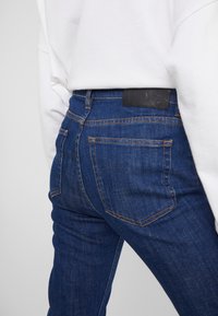 Slim fit tmavě modré džíny z denimu s texturovaným povrchem, oranžovým prošíváním a černou koženou nášivkou na pasu.