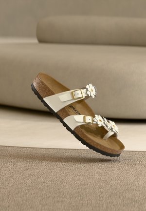Λευκό σανδάλι Birkenstock με δύο ρυθμιζόμενες χρυσές αγκράφες και λευκές διακοσμήσεις μελουσιών σε φελλό σόλας, σε μπεζ φόντο μοκέτας.