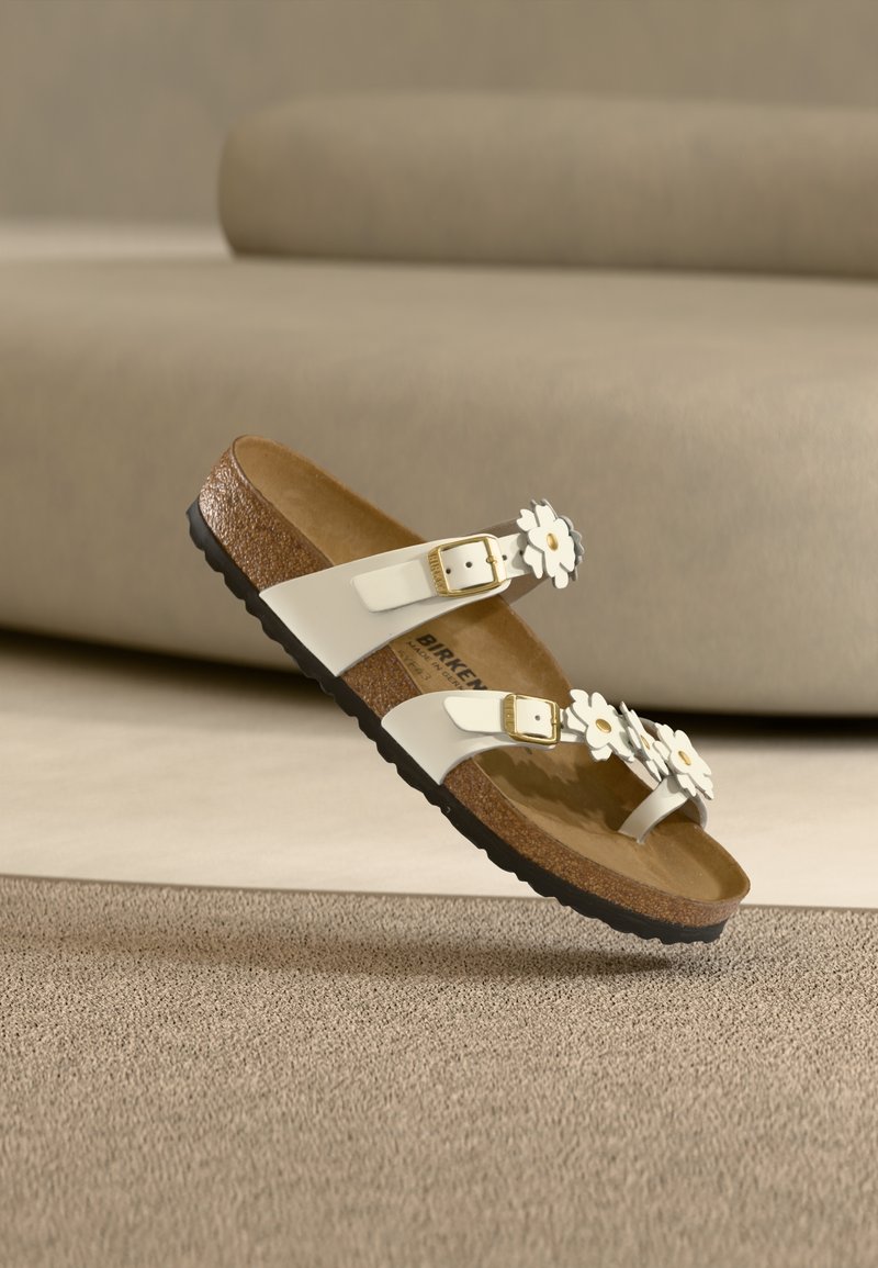 Λευκό σανδάλι Birkenstock με δύο ρυθμιζόμενες χρυσές αγκράφες και λευκές διακοσμήσεις μελουσιών σε φελλό σόλας, σε μπεζ φόντο μοκέτας.