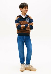 Granatowo-brązowy hoodie w paski z logo, zestawiony z niebieskimi dżinsami i beżowymi trampkami. Ubrania mają luźny krój i casualowy styl.