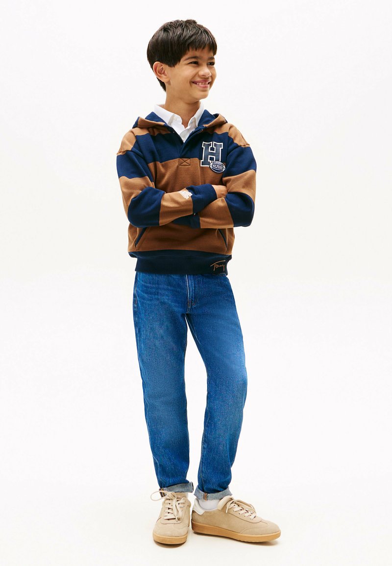 Hoodie rayé bleu marine et marron avec un logo, associé à un jeans en denim bleu et des baskets beiges. Les vêtements ont une coupe décontractée et un style casual.