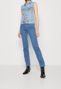 Ljusblå denim ärmlös väst med två bröstfickor, kombinerad med blå skinny jeans och svarta ankelskoblastövlar. Slät textur och figurnära design.