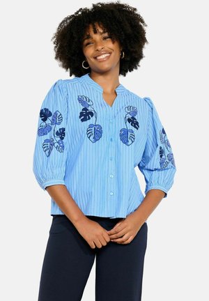 Smiling woman with curly hair wears a blue button-up blouse with vertical stripes and dark blue floral embroidery on sleeves and chest.