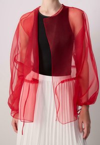 Rote transparente Jacke mit elastischen Details, offenem Vorderteil, weiten Ärmeln und einem Überlagerungsdesign. Kombiniert mit einem schwarzen Top und einem weißen plissierten Rock.