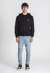 Sudadera con cremallera negra, capucha, bolsillos frontales y cordones. Combinada con unos pantalones vaqueros ajustados azul claro y zapatillas negras con suelas blancas.