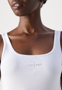 Close-up van een persoon die een witte geribbelde tanktop draagt met een verhoogd Calvin Klein-logo op de borst.