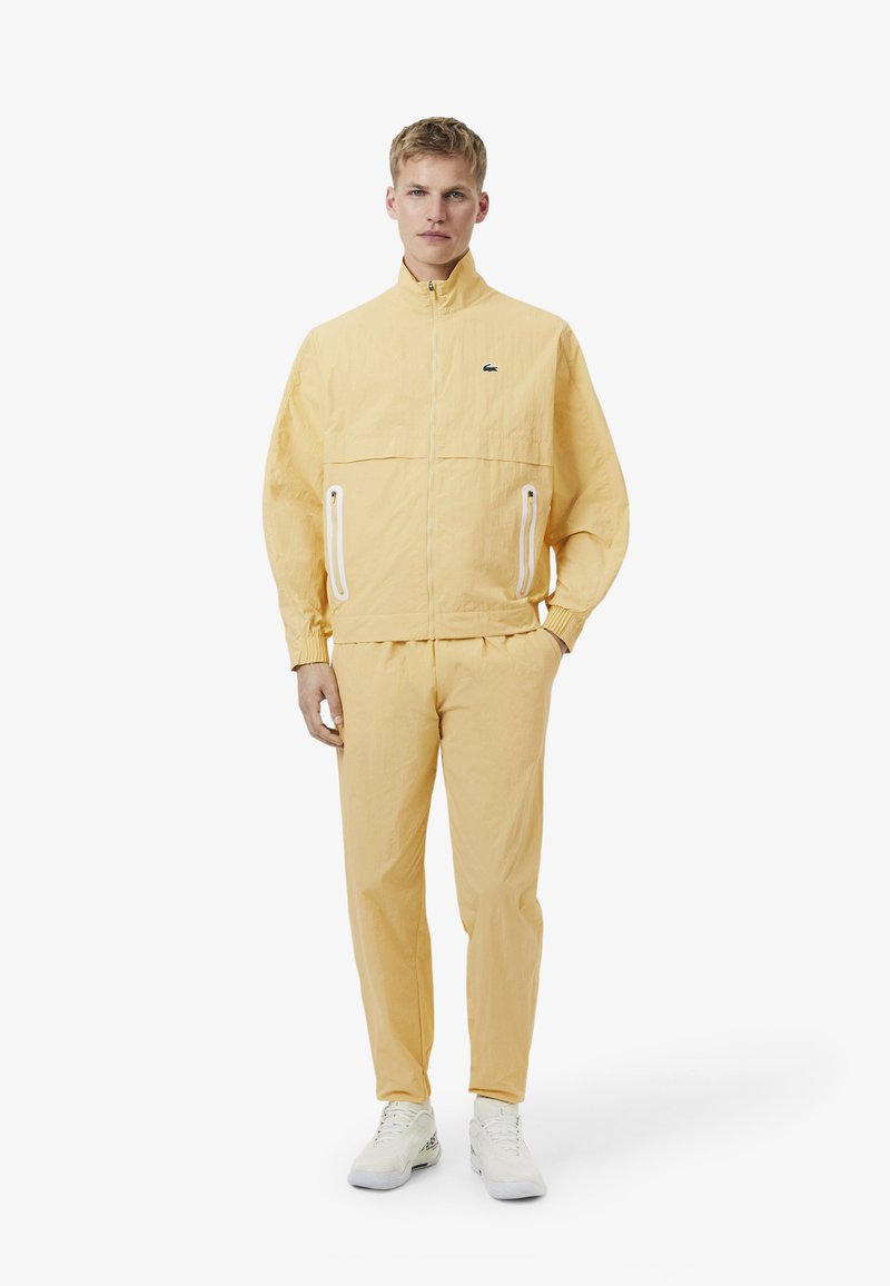 Man met een bleekgele jas met rits en bijpassende broek met witte sneakers, staand tegen een effen witte achtergrond.