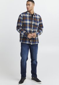 Rutet jakke i blå, brun og kremfarger; knappelukking med en brystlomme; kombinert med mørke jeans og svarte joggesko.