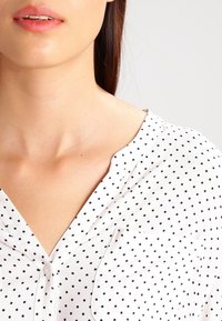 Camicia bianca con collo a V e piccoli pois neri. Tessuto morbido con una tasca sul petto e maniche corte.