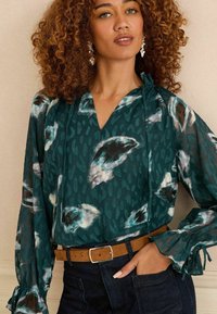 Blusa in teal con motivi astratti bianchi e neri; presenta un collo a V, scollatura arricciata e maniche lunghe con polsini arricciati.