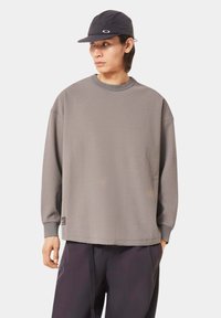 Graues, strukturiertes Langarmshirt mit Rundhalsausschnitt. Verfügt über gerippten Stoff und passende Ärmelbündchen. Getragen mit einer dunklen Kappe, die den lässigen Stil betont.