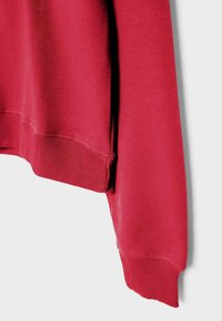 Manica e orlo di felpa rossa con polsini a costine su sfondo bianco, che mostrano la trama del tessuto e i dettagli delle cuciture.