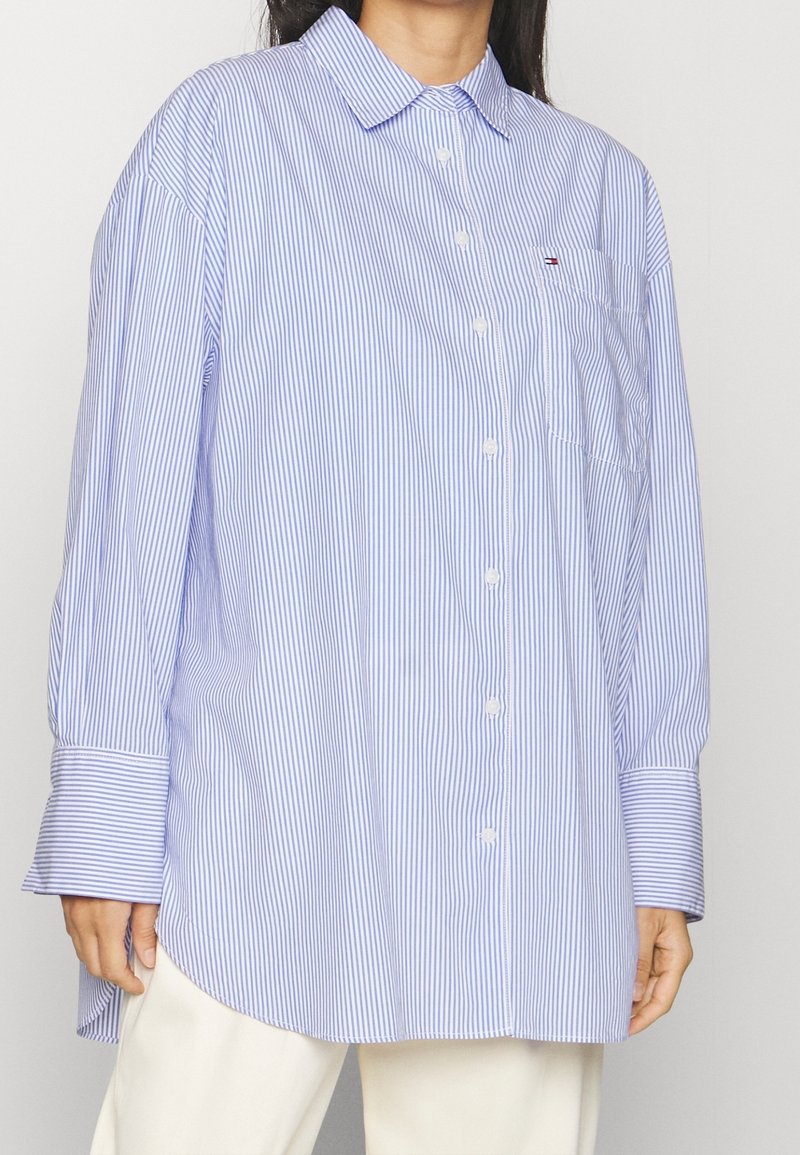 Femme portant une chemise à rayures verticales bleues et blanches oversize avec une poche poitrine et un pantalon blanc.