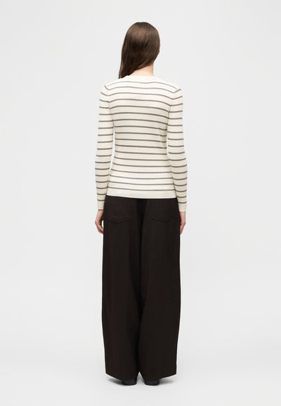Γυναίκα με μακριά καστανά μαλλιά στέκεται με την πλάτη γυρισμένη φορώντας μια κρεμ πλεκτή μπλούζα με λεπτές καφέ ρίγες και φαρδιά μαύρη παντελόνια με πίσω τσέπες.