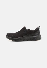 Zapatillas negras sin cordones hechas de malla transpirable, con una suela flexible y acentos sutiles. Diseño simple sin patrones visibles ni elementos de ferretería.
