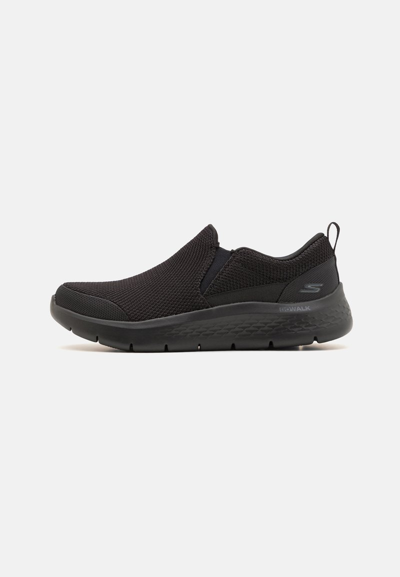 Zapatillas negras sin cordones hechas de malla transpirable, con una suela flexible y acentos sutiles. Diseño simple sin patrones visibles ni elementos de ferretería.