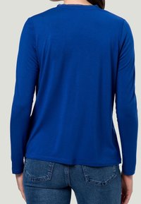 Donna che indossa una semplice maglietta a maniche lunghe blu royal e jeans in denim blu, vista da dietro su uno sfondo grigio chiaro.