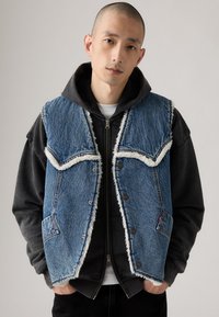 Denimvest med pelsfor, blå farve, frynset kanter og kontrasterende lommer. Båret over en sort hættetrøje med lynlåse og detaljer.