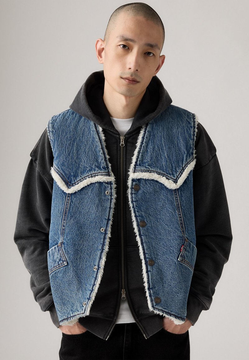Denimvest med pelsfor, blå farve, frynset kanter og kontrasterende lommer. Båret over en sort hættetrøje med lynlåse og detaljer.