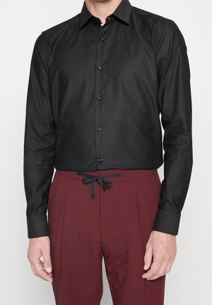 Mann trägt ein eng anliegendes schwarzes Hemd mit Knöpfen und bordeauxrote Hose mit Kordelzug, steht mit entspannt an den Seiten hängenden Händen.