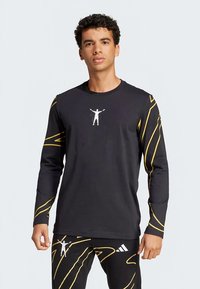 Chemise noire à manches longues avec des lignes abstraites jaunes, arborant un graphique blanc d'une silhouette aux bras levés sur le devant. Texture lisse.