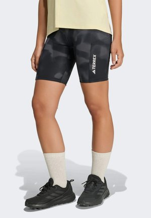 Benen met zwarte Adidas Terrex shorts met camouflering patroon, crèmekleurige sokken en zwarte Adidas sneakers op een lichtgrijze achtergrond.