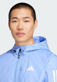 Veste de course bleu clair avec capuche, fermeture éclair sur le devant et accents de logo blanc sur la poitrine et les manches. Matériau lisse et léger.