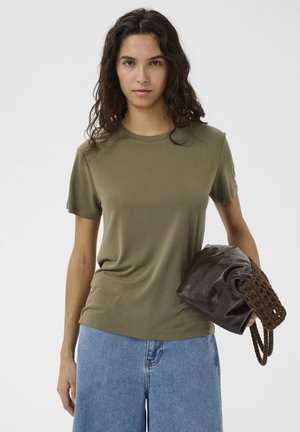 Femme aux longs cheveux foncés porte un t-shirt vert olive et un jean bleu, tenant un sac à main en cuir marron et tissé sur un fond uni.