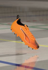 Oranžid jalgpalliketsad, millel on voolujooneline disain, tekstuuriga ülemine osa, must sisemus ja hõbedased detailid. Varustatud vormitud naeladega haardeks.