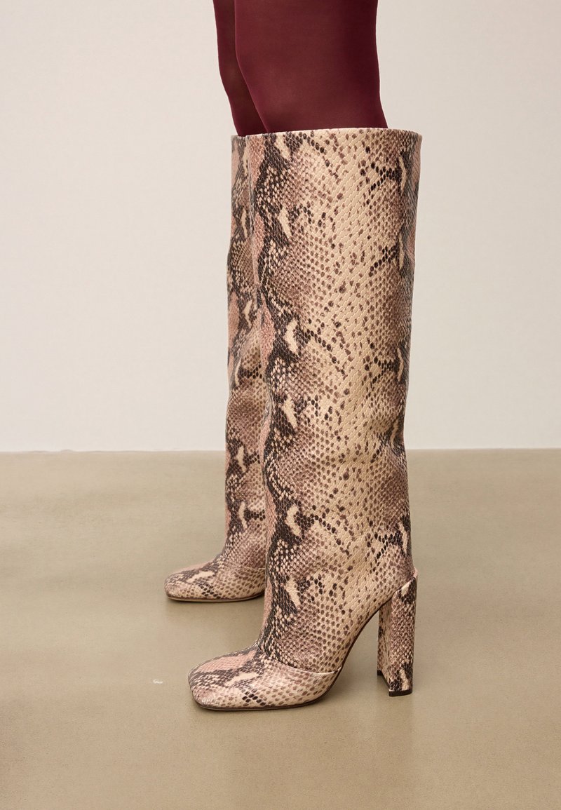 Bottes à talons hautes, arrivant au genou, avec un motif en serpent beige et marron portées avec des collants bordeaux foncé sur un sol lisse.