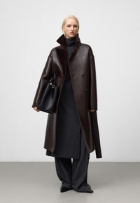 Cappotto in pelle marrone con colletto alto e finitura texturizzata; abbinato a un vestito grigio scuro e pantaloni a gamba larga; con una borsa nera.