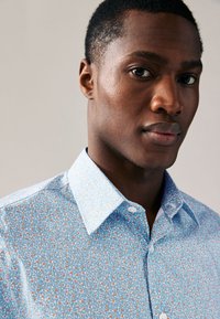 Ljusblå skjorta med knappar, prydd med ett blommönster i orange och vitt, med en strukturerad krage och mjuk tygstruktur.