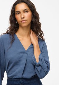 Blaue, strukturierte Bluse mit V-Ausschnitt, langen Ärmeln und Knopfmanschetten, kombiniert mit einer dunklen Denim-Hose.