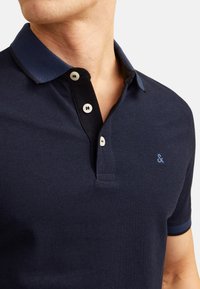 Marineblauwe poloshirt van een gestructureerde stof, met een contrasterende kraag in donkerblauw, een drie-knopen sluiting en een geborduurd logo op de borst.