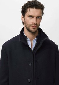 Abrigo de lana negro con un alto cuello, cierre de botones y bolsillos laterales. Llevado sobre una camisa a rayas azul claro y blanca.