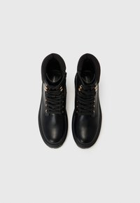 Chaussures montantes en cuir noir avec un col de cheville côtelé, des lacets noirs, des œillets dorés et un bout lisse et arrondi. Marque discrète à l'intérieur.